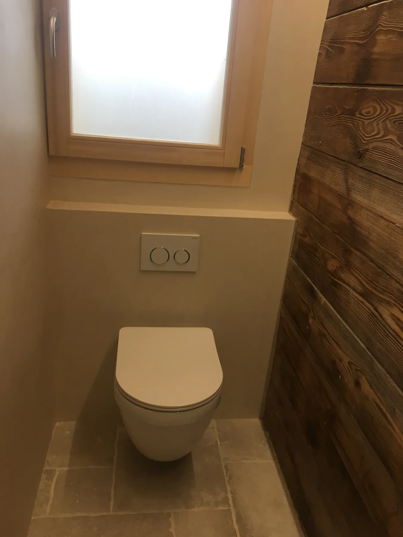 Des toilettes suspendues modernes avec un mur en bois habillé et un carrelage au sol, sous une fenêtre en bois dépolie — chantier Bio Rénovation, Chablais