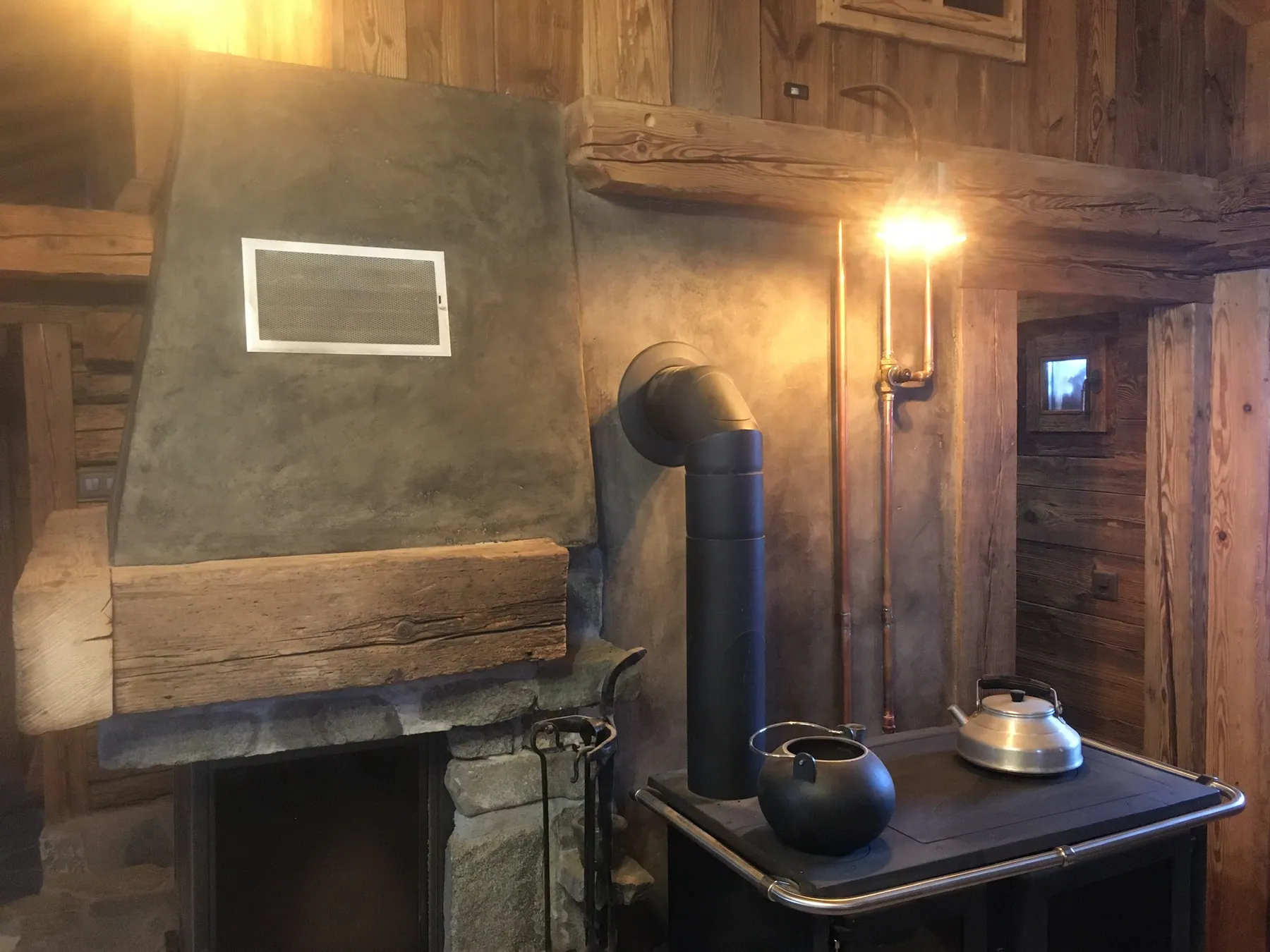 Un intérieur chaleureux de style chalet avec une cheminée en pierre et microciment, des murs en bois, et un poêle à bois avec sa tuyauterie en cuivre apparente — chantier Bio Rénovation, Alpes vaudoises
