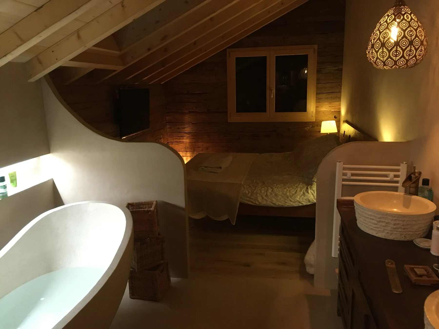 Une chambre mansardée chaleureuse avec un lit double et un espace salle de bains ouvert, éclairée par des suspensions décoratives — chantier Bio Rénovation, Alpes vaudoises