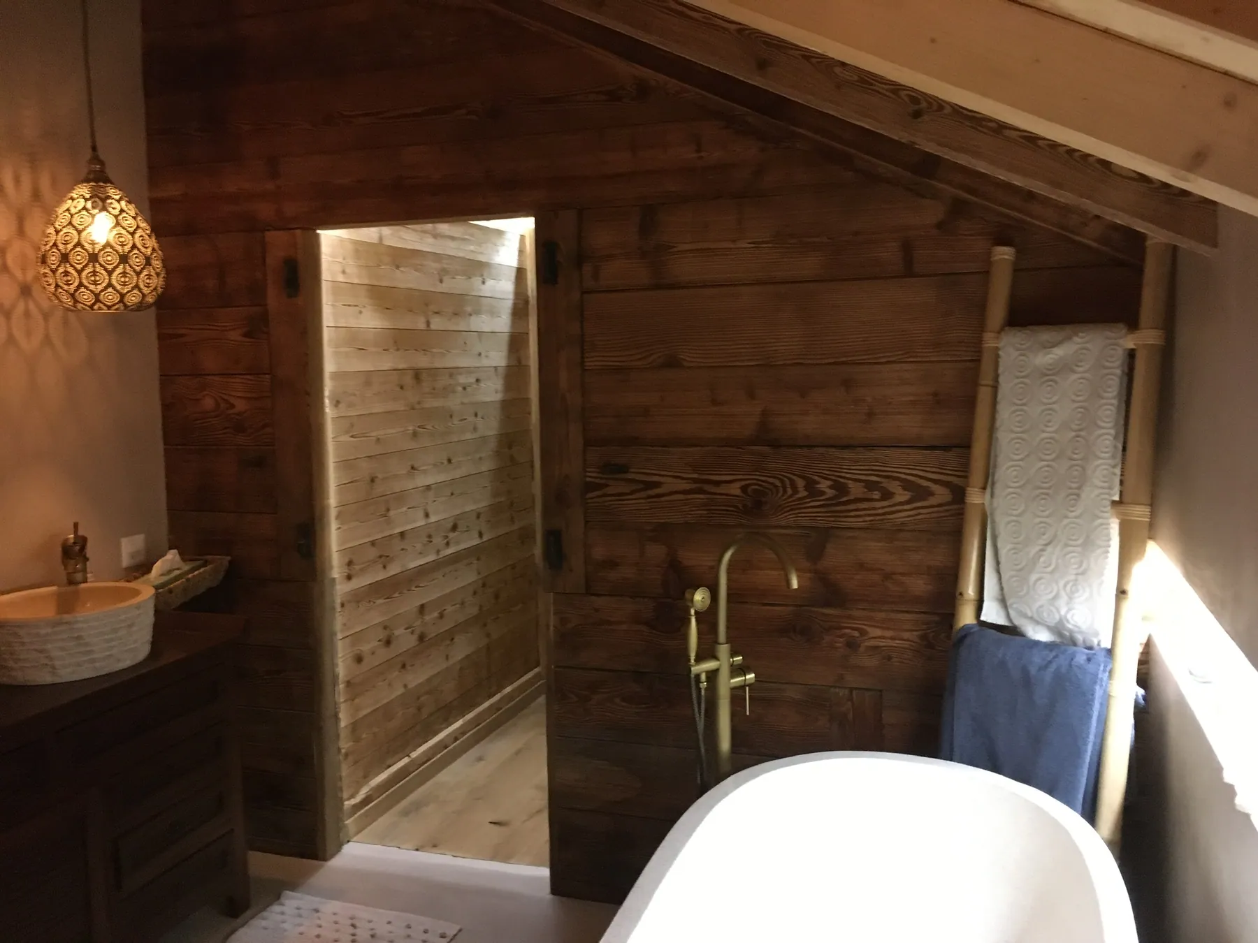 Une salle de bain mansardée au style chalet avec des murs en bois, une vasque en pierre et une baignoire blanche, agrémentée d'une robinetterie dorée et d'un porte-serviettes en bambou — chantier Bio Rénovation, Val de Bagnes / Verbier
