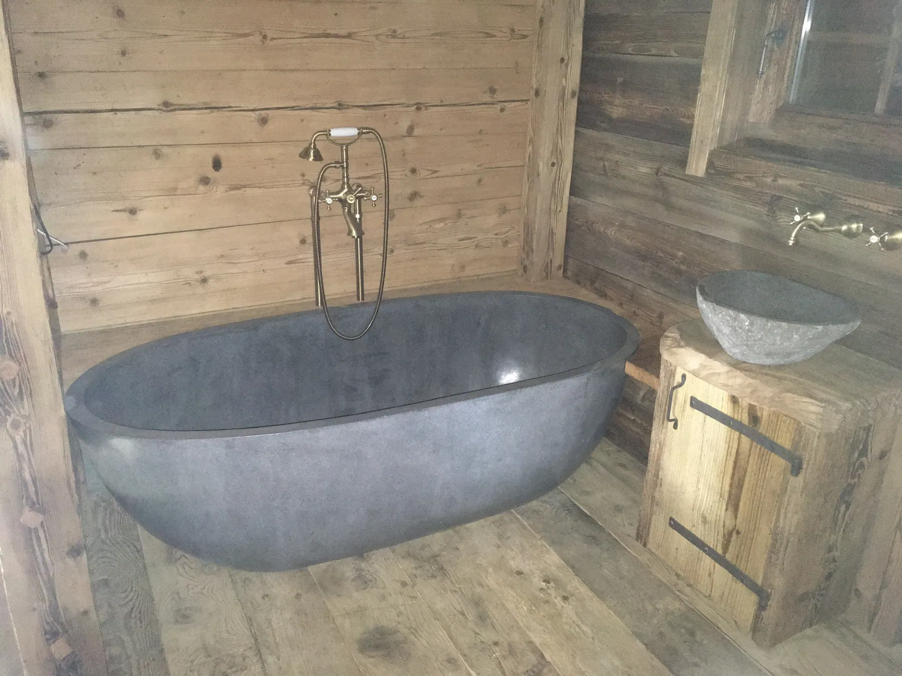 Une salle de bain rustique avec des murs et un sol en planches de bois brut, une baignoire autoportante grise de style ancien et une vasque en pierre posée sur un meuble en bois — chantier Bio Rénovation, Val de Bagnes / Verbier