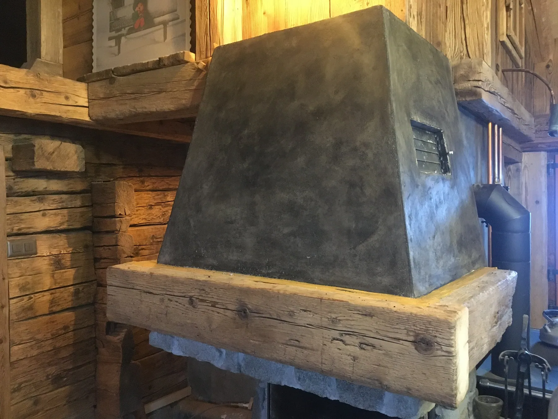 Un foyer de cheminée moderne en béton ciré, avec un insert, et son manteau en bois massif dans un intérieur de chalet — chantier Bio Rénovation, Alpes vaudoises