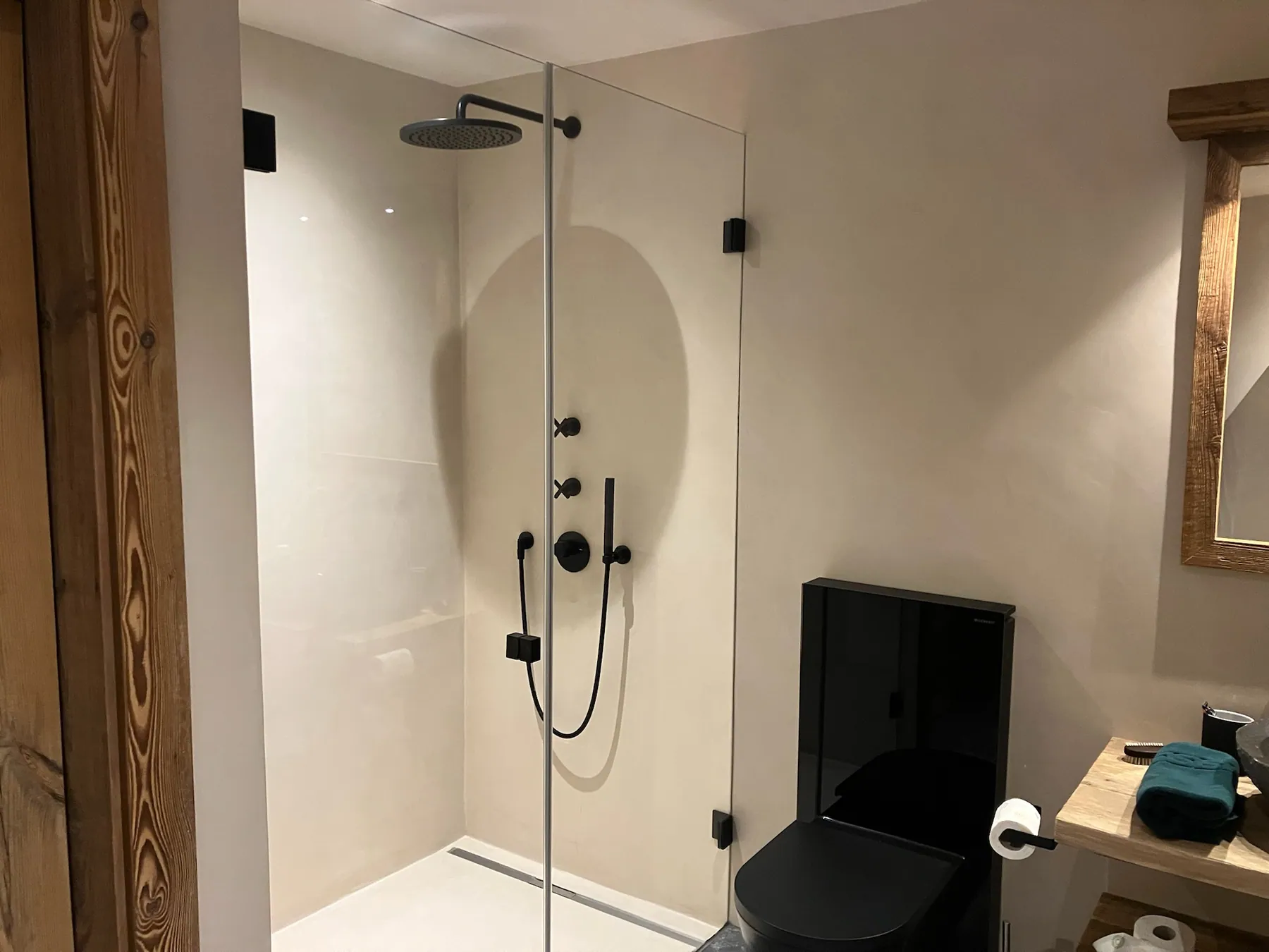 Une salle de bains au design contemporain avec une grande douche vitrée et des éléments noirs, un WC noir et un meuble vasque en bois rustique — chantier Bio Rénovation, Villars-Gryon (VD)