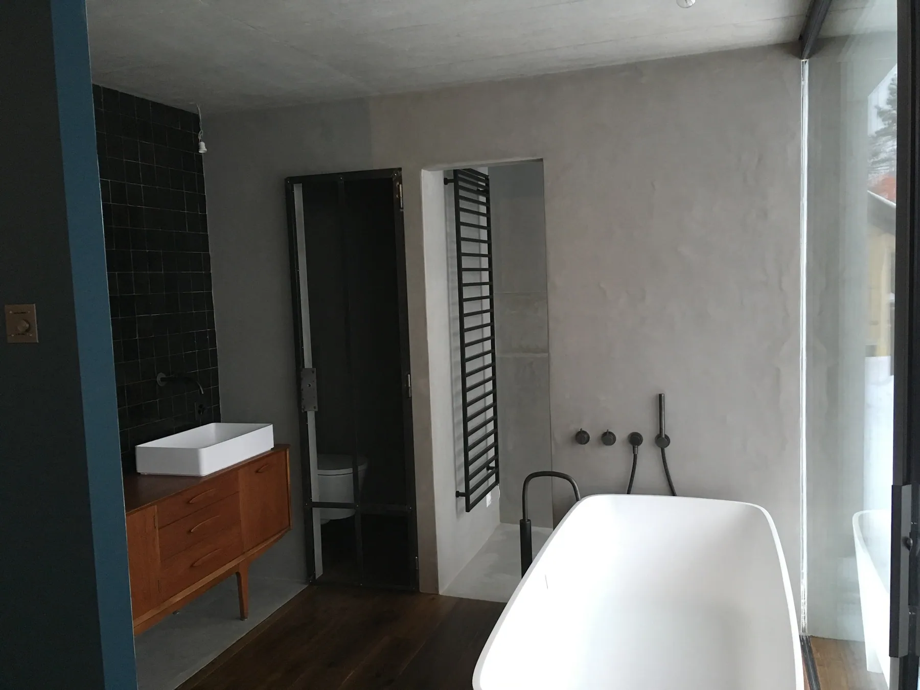 Une salle de bain moderne avec des murs en béton ciré, un meuble sous vasque en bois foncé et une baignoire blanche, complétée par une paroi de douche vitrée et un carrelage mural sombre — chantier Bio Rénovation, Suisse romande