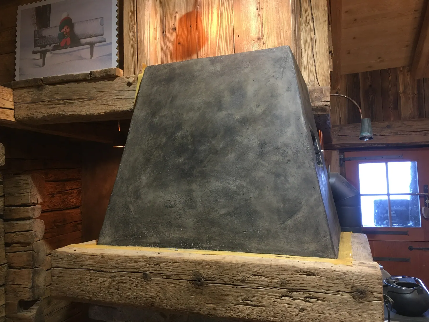 Une hotte de cheminée en béton ciré gris surmontant une poutre en bois massif dans un intérieur de style chalet — chantier Bio Rénovation, Alpes vaudoises