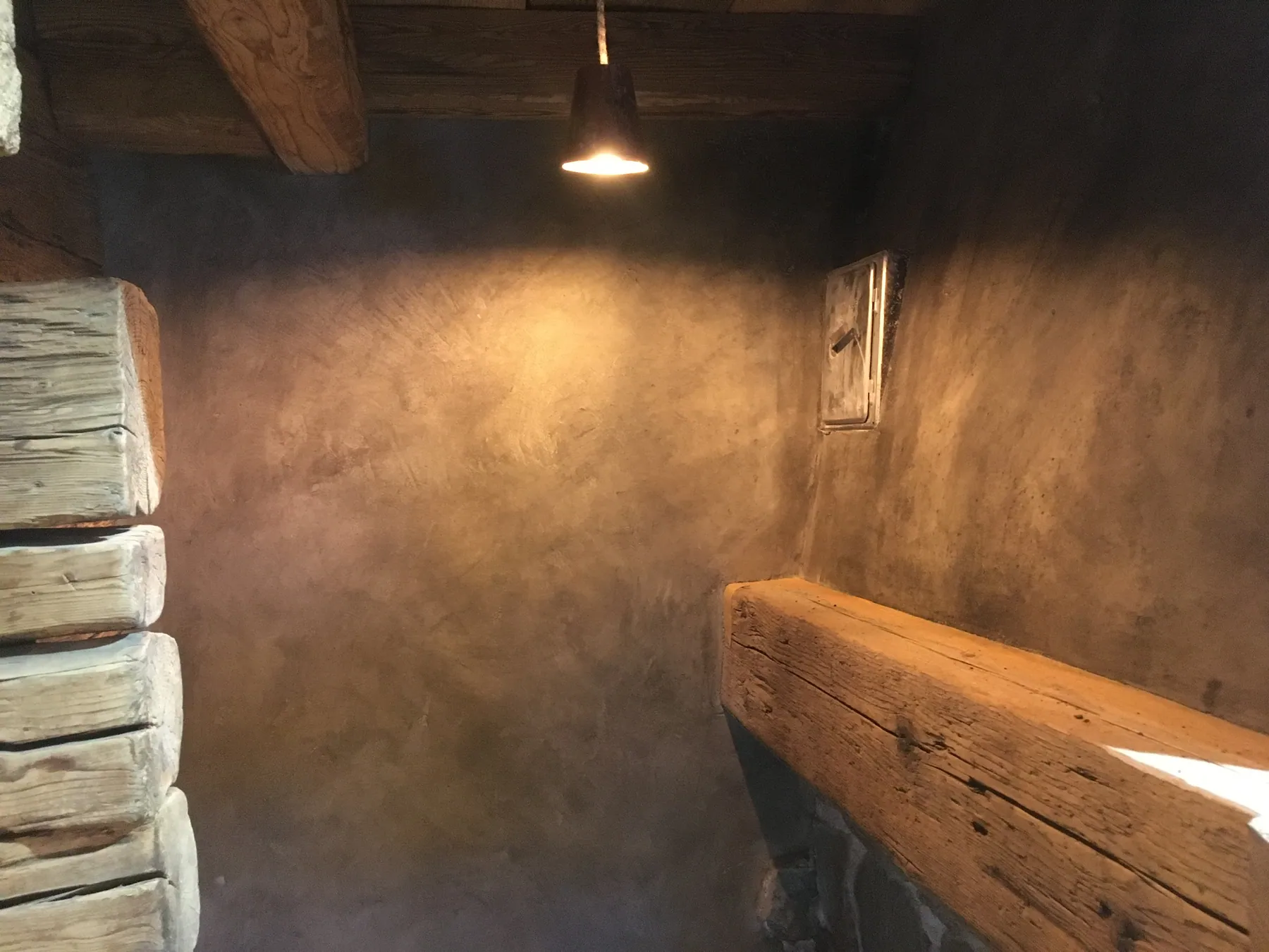 Un mur en béton ciré sombre avec une applique lumineuse contraste avec les poutres en bois apparentes — chantier Bio Rénovation, Alpes vaudoises