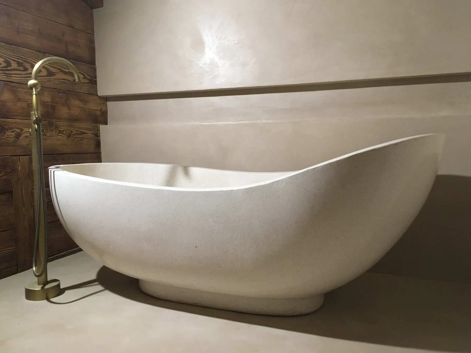 Une baignoire autoportante élégante en forme de bateau, positionnée sur un sol en béton ciré devant un mur en planches de bois rustiques et un mur enduit clair — chantier Bio Rénovation, Riviera vaudoise