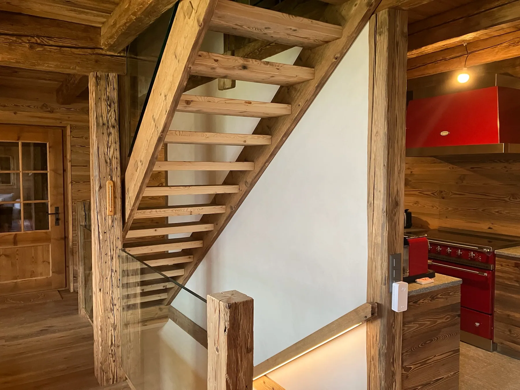 Un escalier en bois massif avec des garde-corps en verre, intégrant une cuisine moderne avec des éléments rouges — chantier Bio Rénovation, Villars-Gryon (VD)