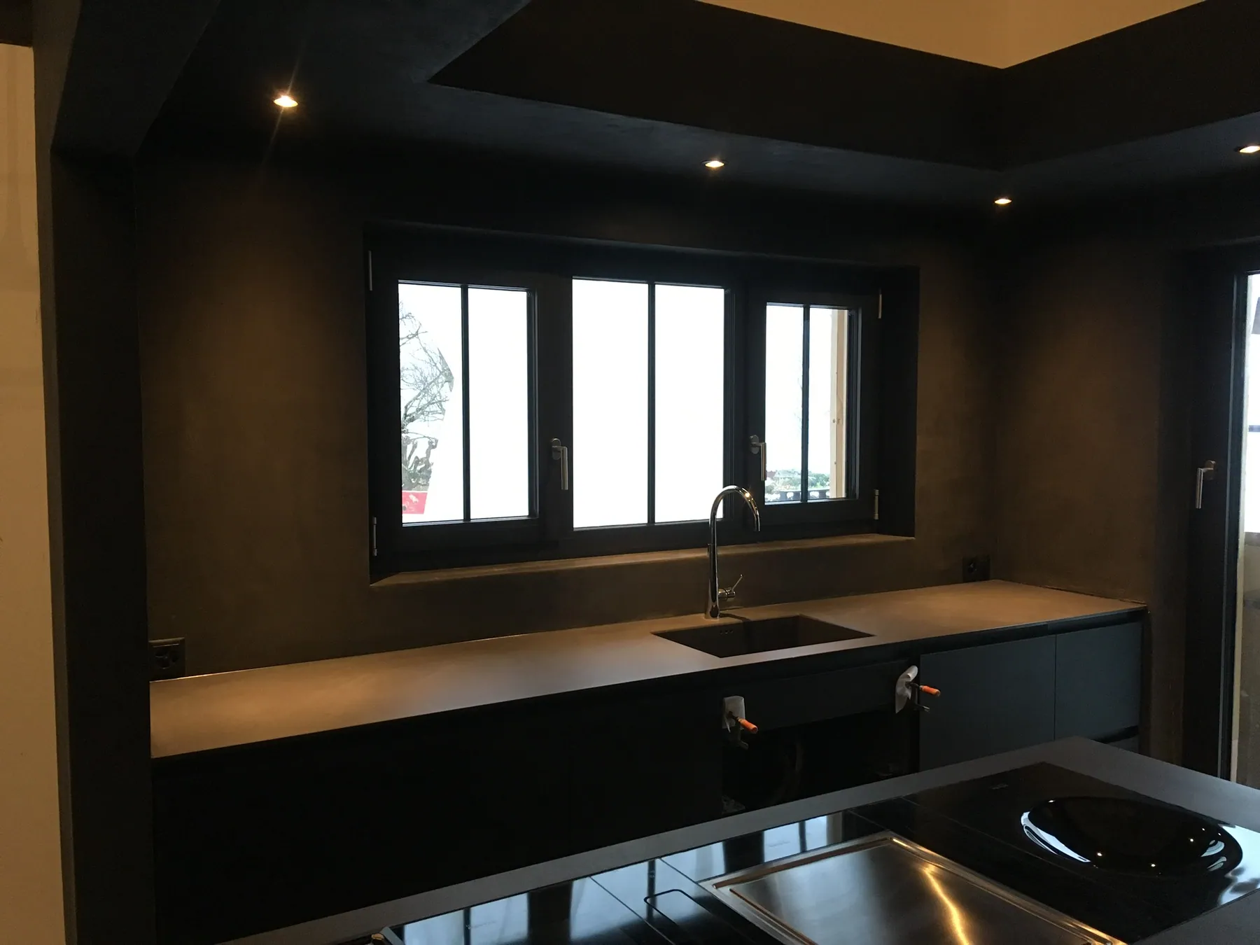 Une cuisine moderne et sombre avec des murs en béton ciré anthracite, des meubles bas noirs et un évier sous une fenêtre à petits carreaux — chantier Bio Rénovation, Suisse romande