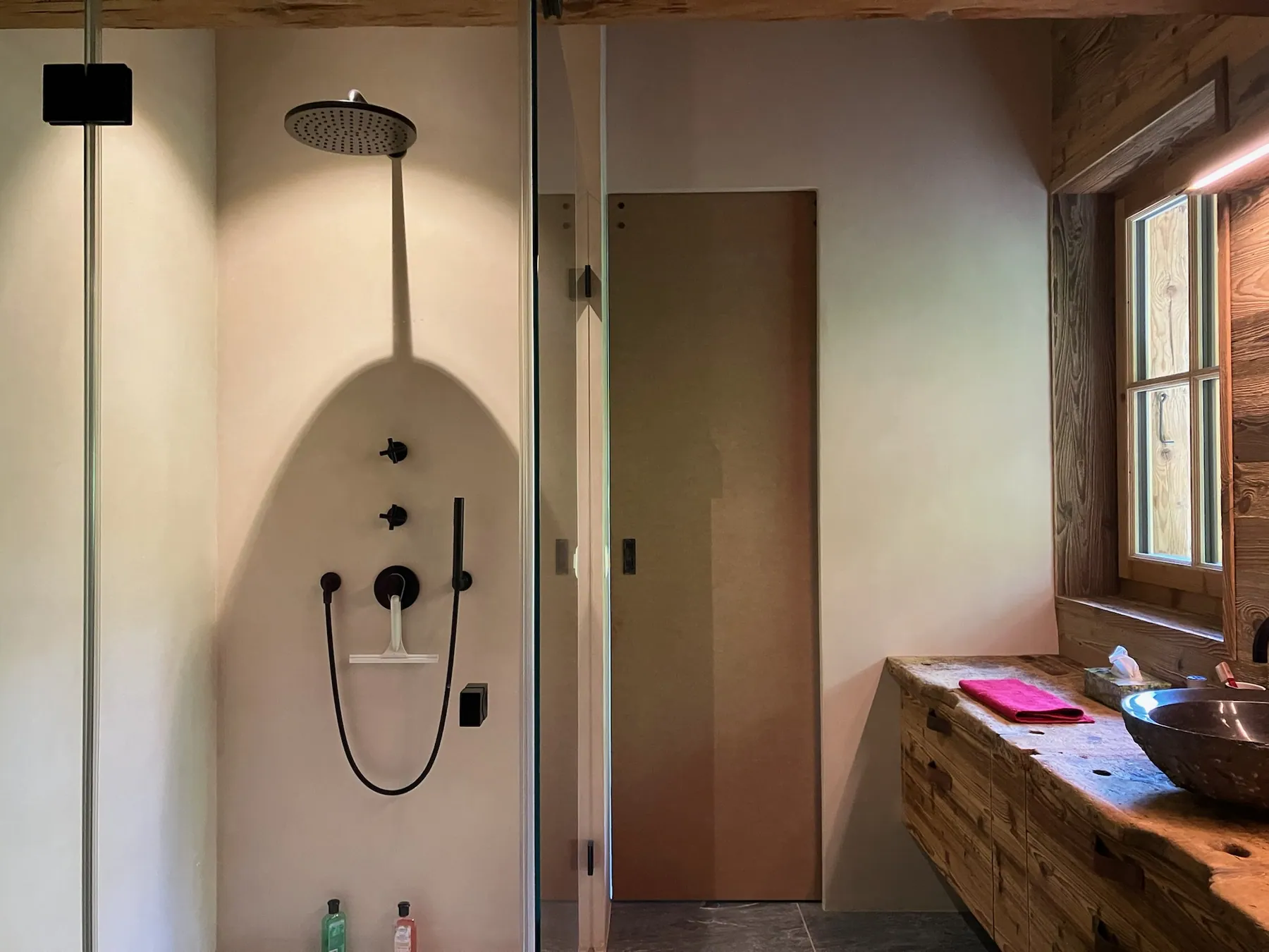 Une salle de bains lumineuse présente une douche à l'italienne et un meuble vasque en bois avec un mur en béton ciré clair — chantier Bio Rénovation, Villars-Gryon (VD)