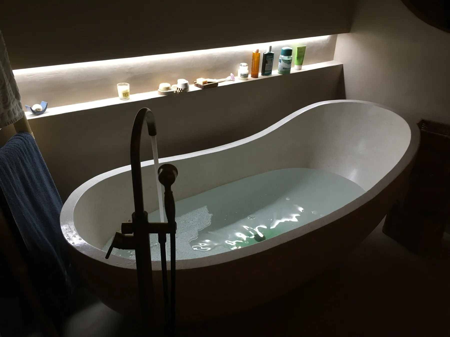 Une baignoire îlot remplie d'eau dans une salle de bains cosy avec un éclairage d'ambiance et des produits sur une étagère intégrée — chantier Bio Rénovation, Alpes vaudoises