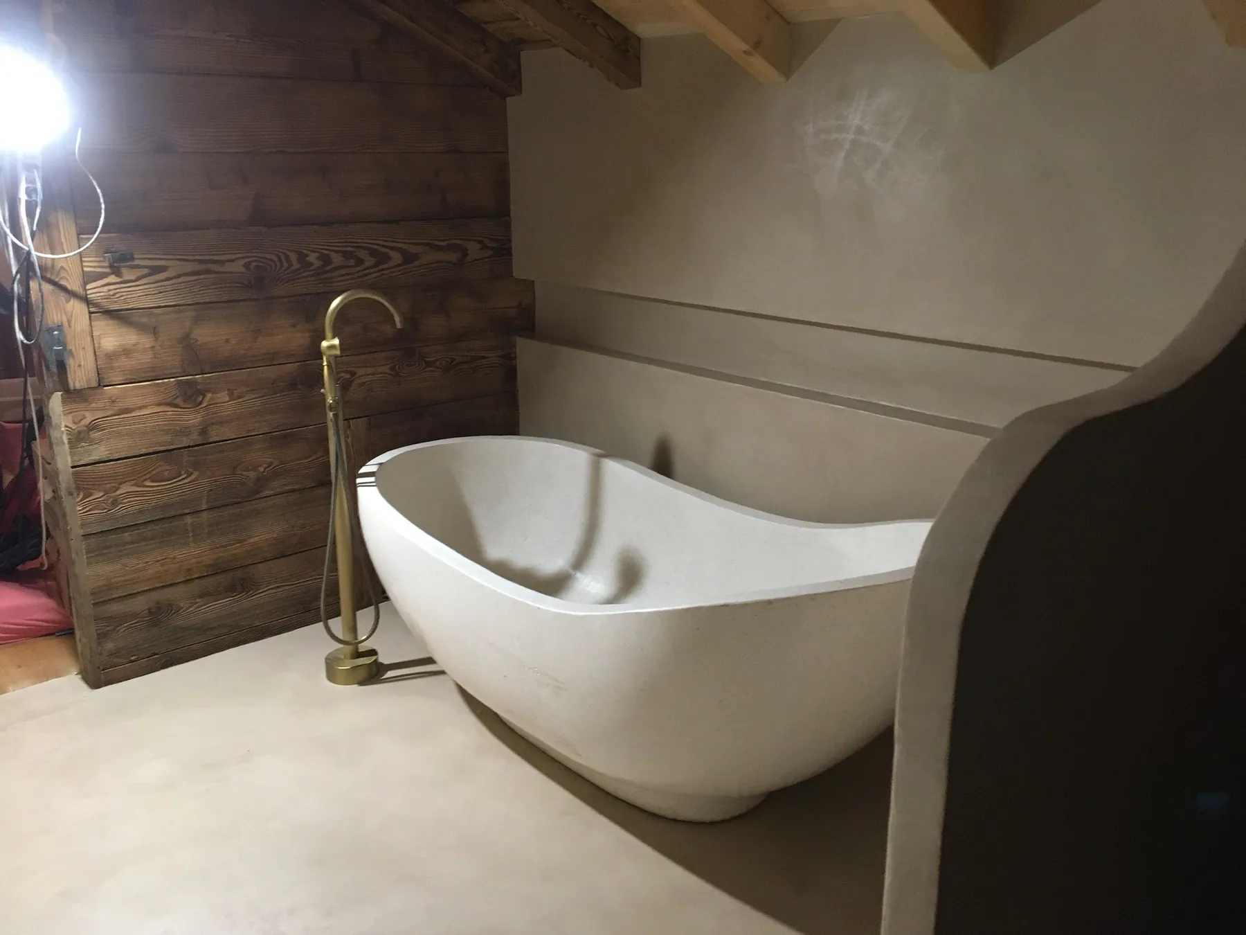 Une baignoire îlot au design moderne avec robinetterie en laiton doré, dans une salle de bains aux murs et sol en béton ciré et un mur en bois — chantier Bio Rénovation, Riviera vaudoise