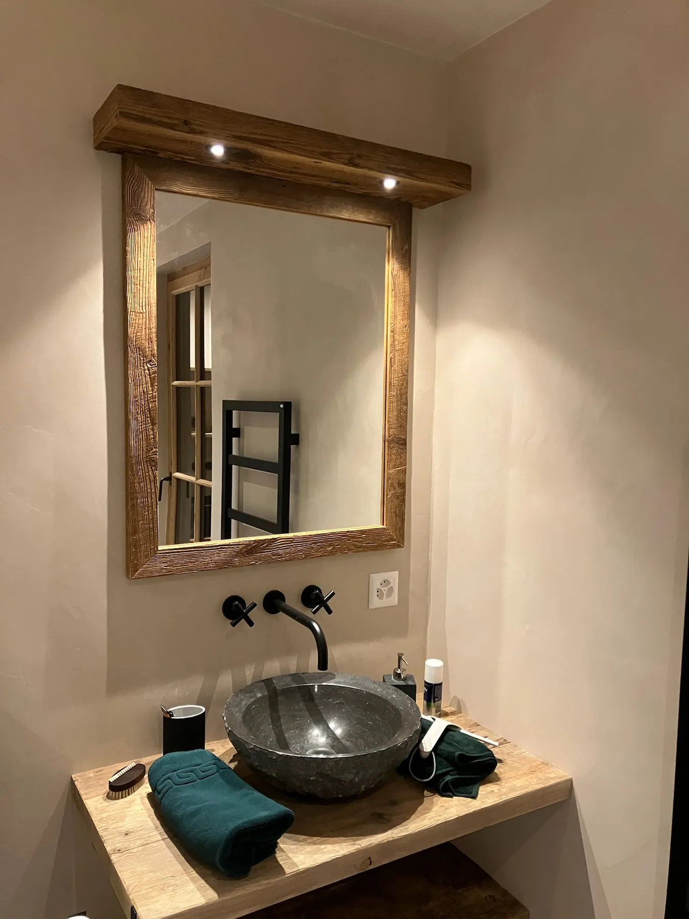Une salle de bains avec un meuble vasque en bois brut, une vasque en pierre noire et un miroir encadré de bois, éclairé — chantier Bio Rénovation, Villars-Gryon (VD)