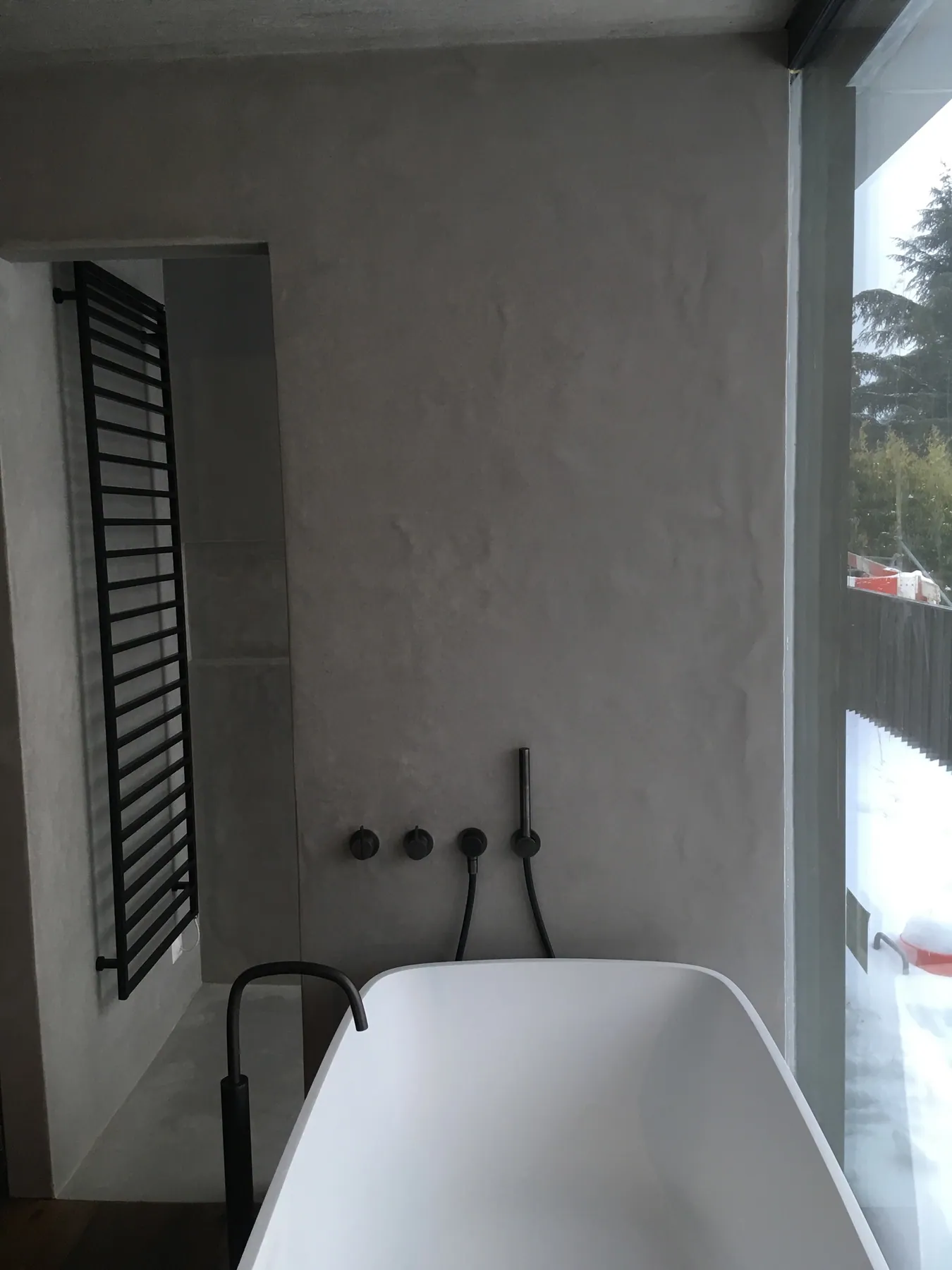 Une salle de bains moderne avec des murs en béton ciré gris, une baignoire îlot blanche et un radiateur sèche-serviettes noir — chantier Bio Rénovation, Suisse romande