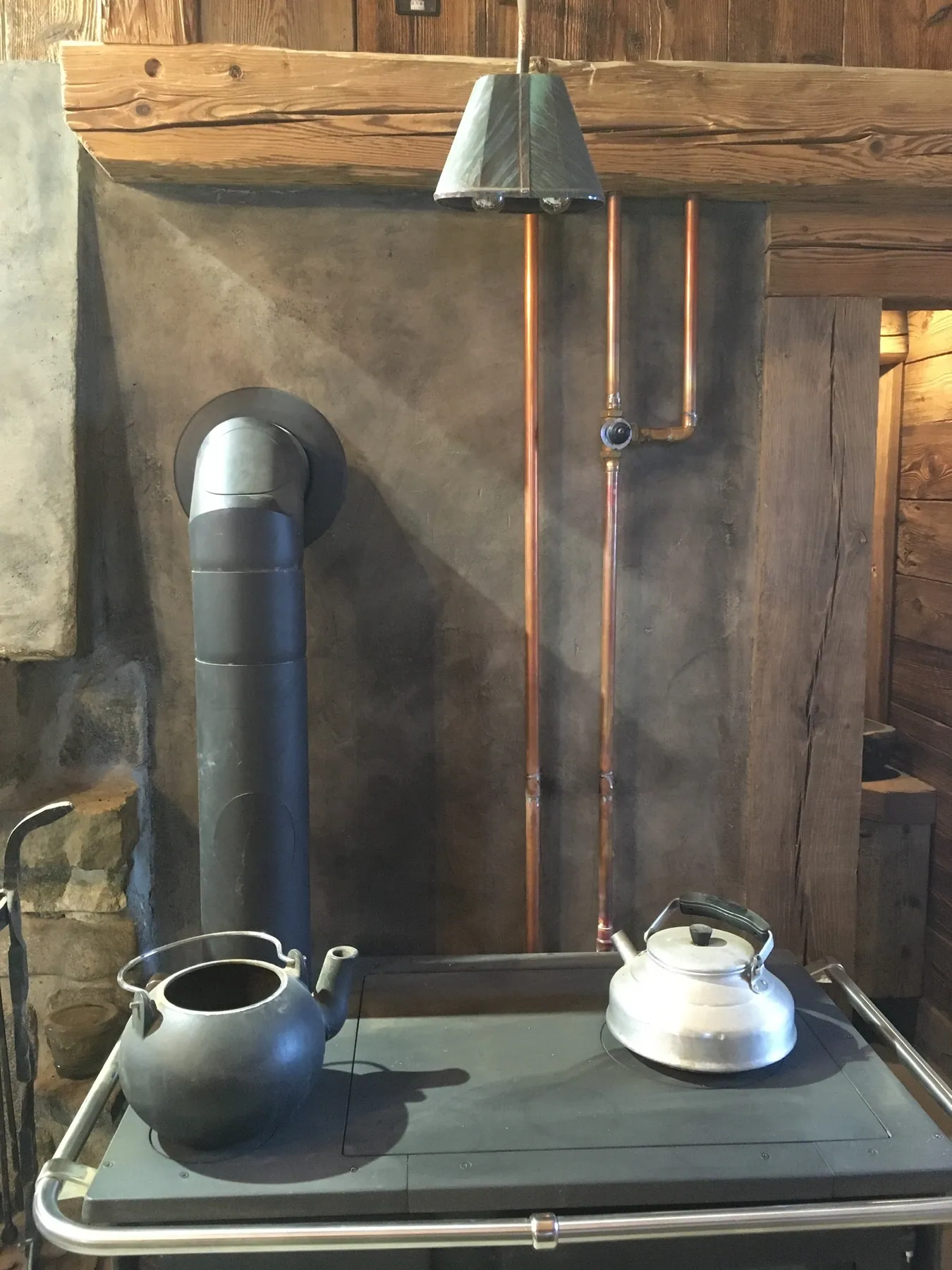 Un poêle de cuisine avec bouilloires posé devant un mur en béton ciré orné de tuyauterie en cuivre apparente et des poutres en bois — chantier Bio Rénovation, Alpes vaudoises