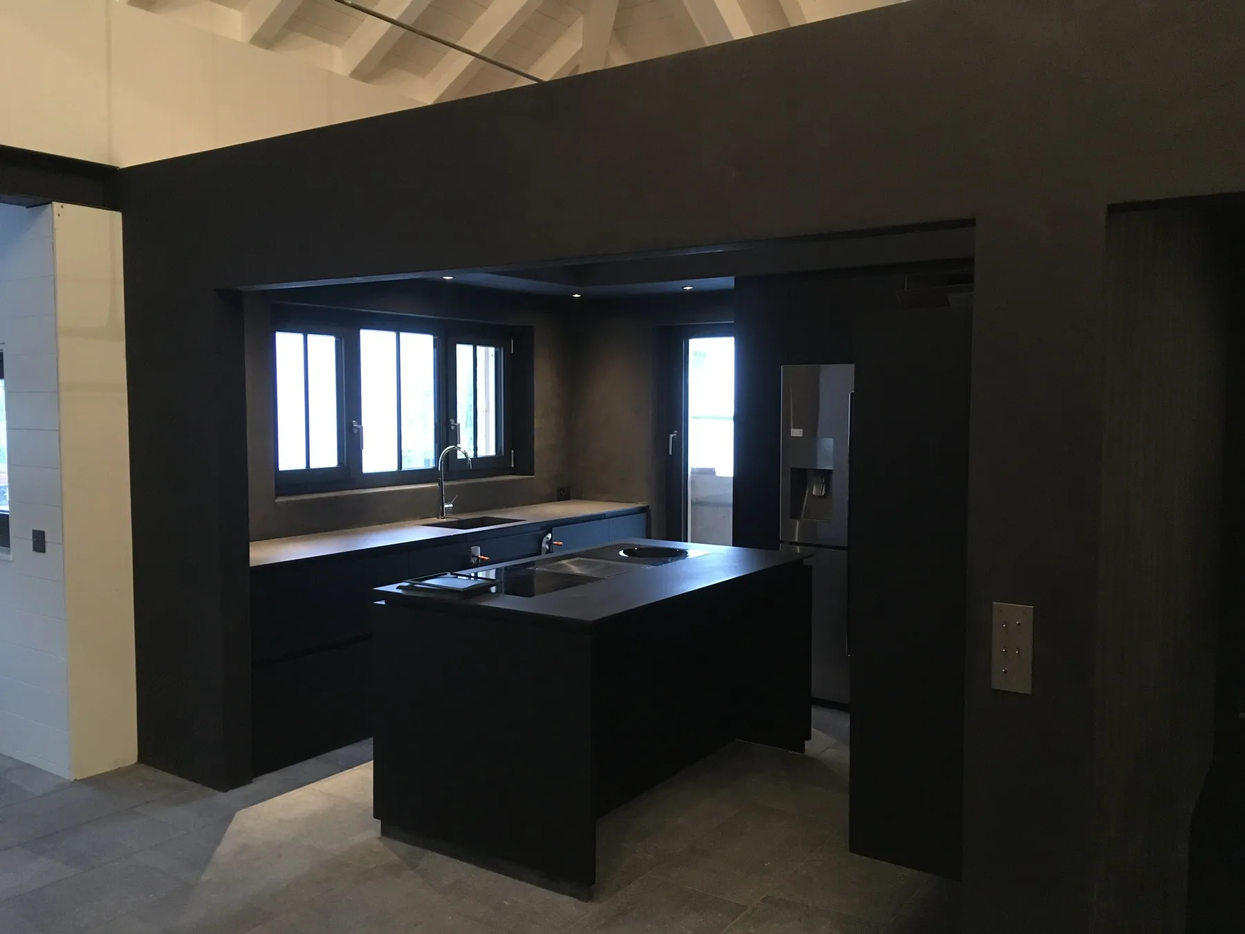 Une cuisine moderne noire avec un îlot central et des éléments en bois, sur un sol carrelé, sous un plafond aux poutres apparentes — chantier Bio Rénovation, Suisse romande